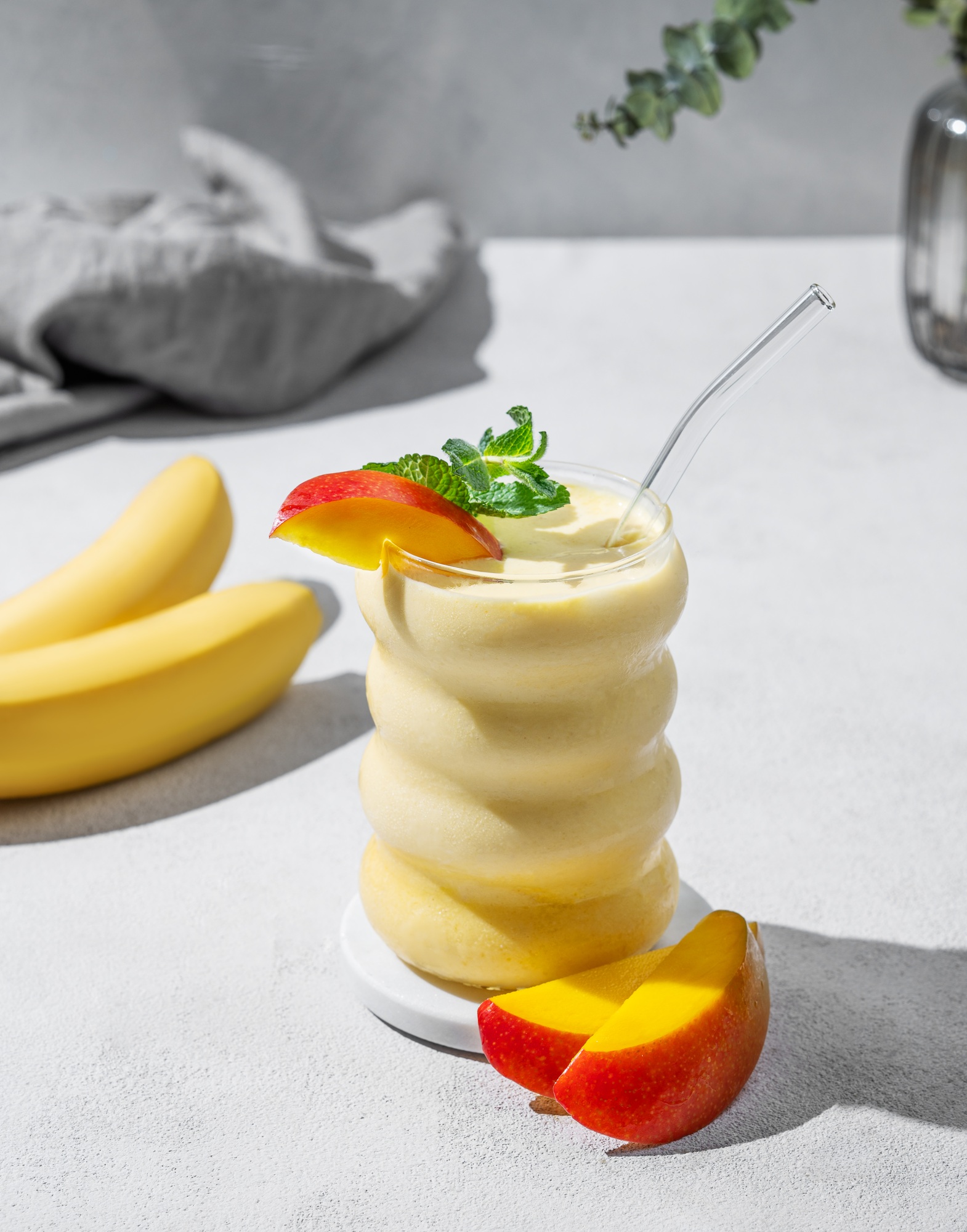 Frozen smoothie with mango and banana in a glass on a light background with tropical fruit slices.